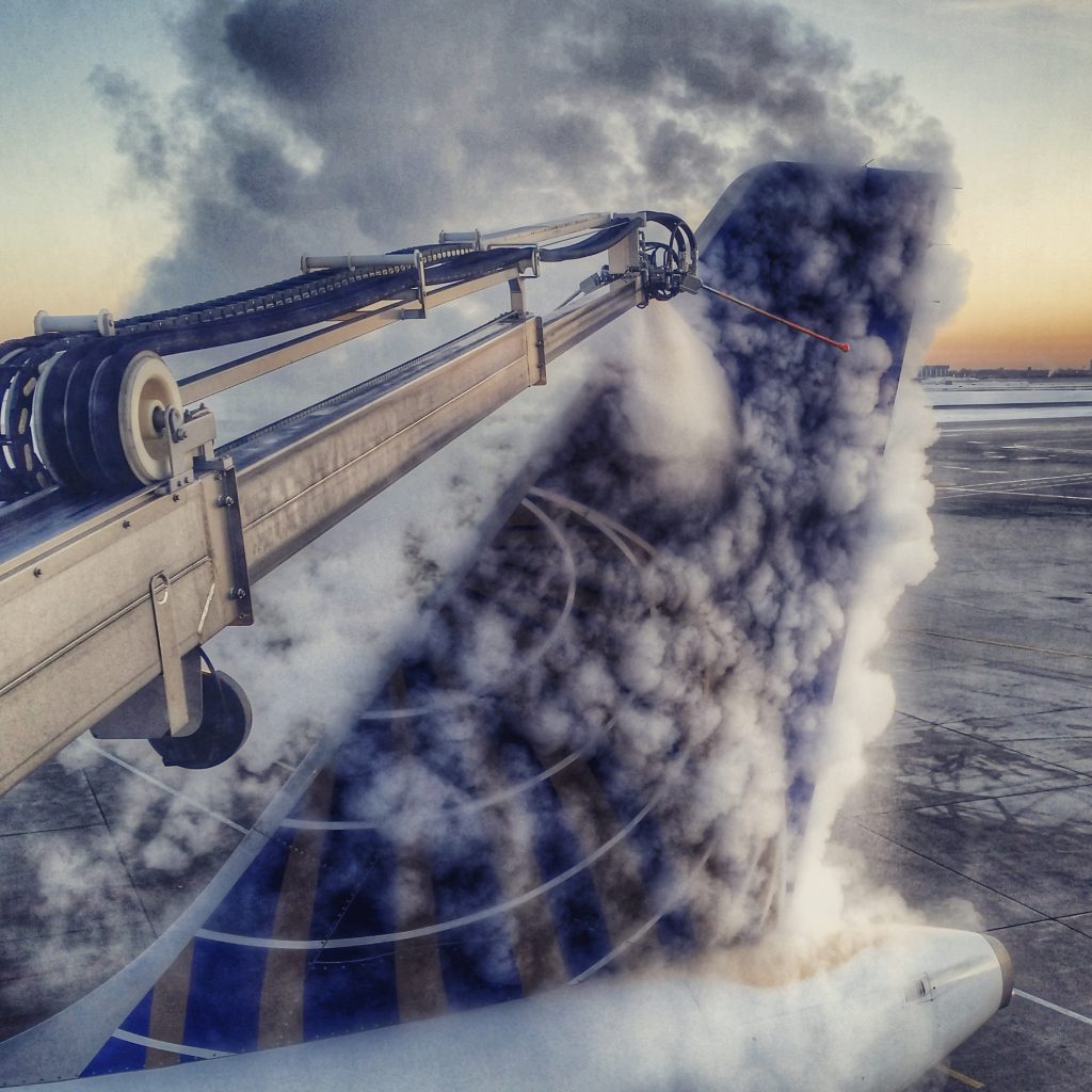 De-icing equipment spraying fluid on an airplane tail, creating a cloud of vapor against a sunset backdrop.