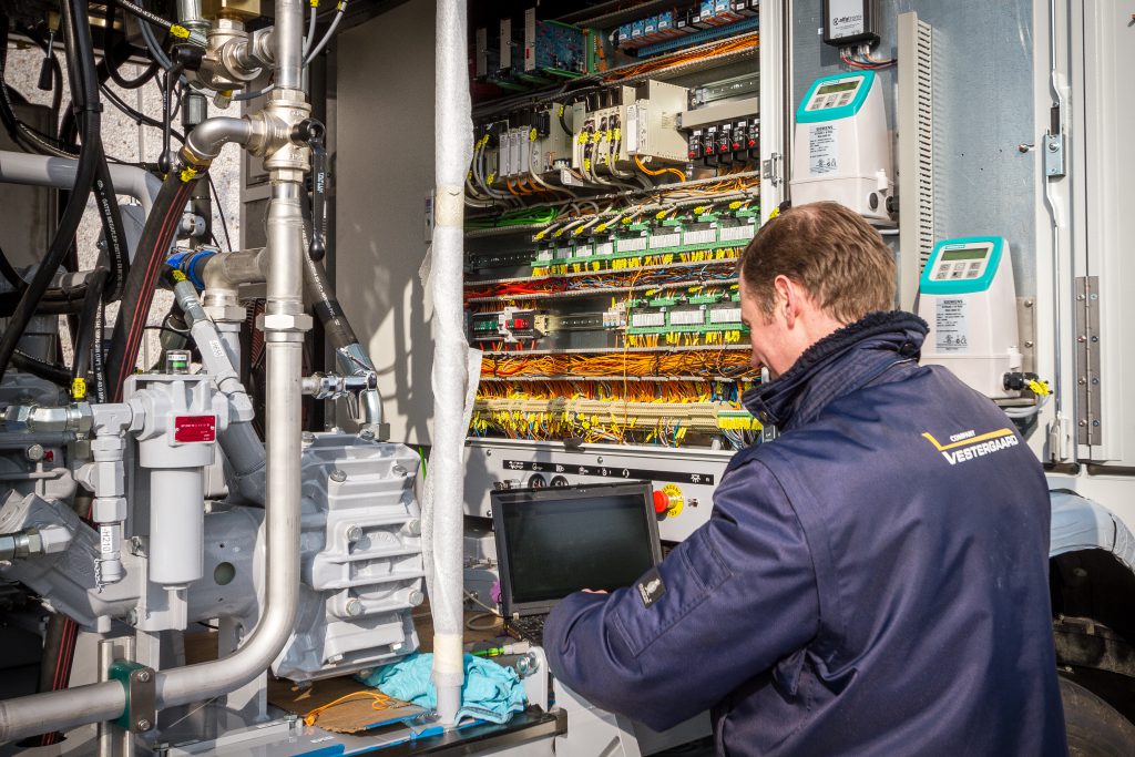 Technician working on electrical equipment with visible wires and a monitor displaying data in a control panel.