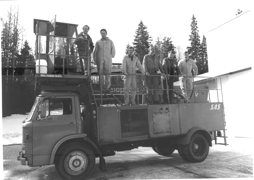 Seven individuals in work overalls stand on a platform atop a vintage truck, surrounded by trees and a building.