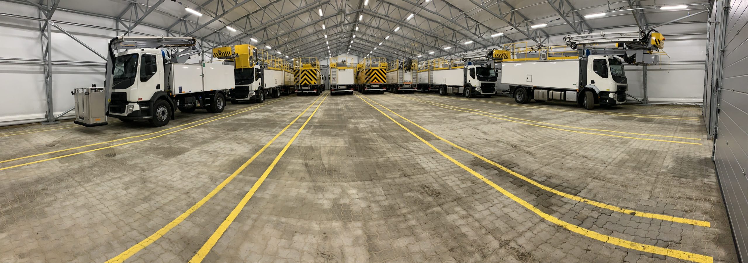 Large indoor space with multiple trucks parked, featuring yellow overhead cranes and marked lanes on the floor.