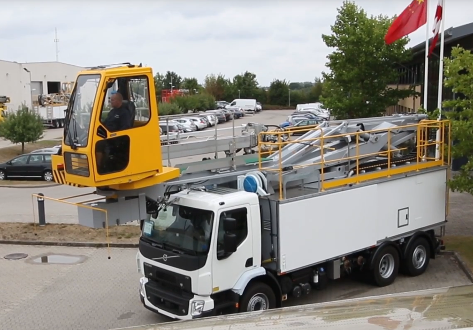 A yellow elevated work platform with an operator above a white truck, parked in an industrial area with trees and flags.