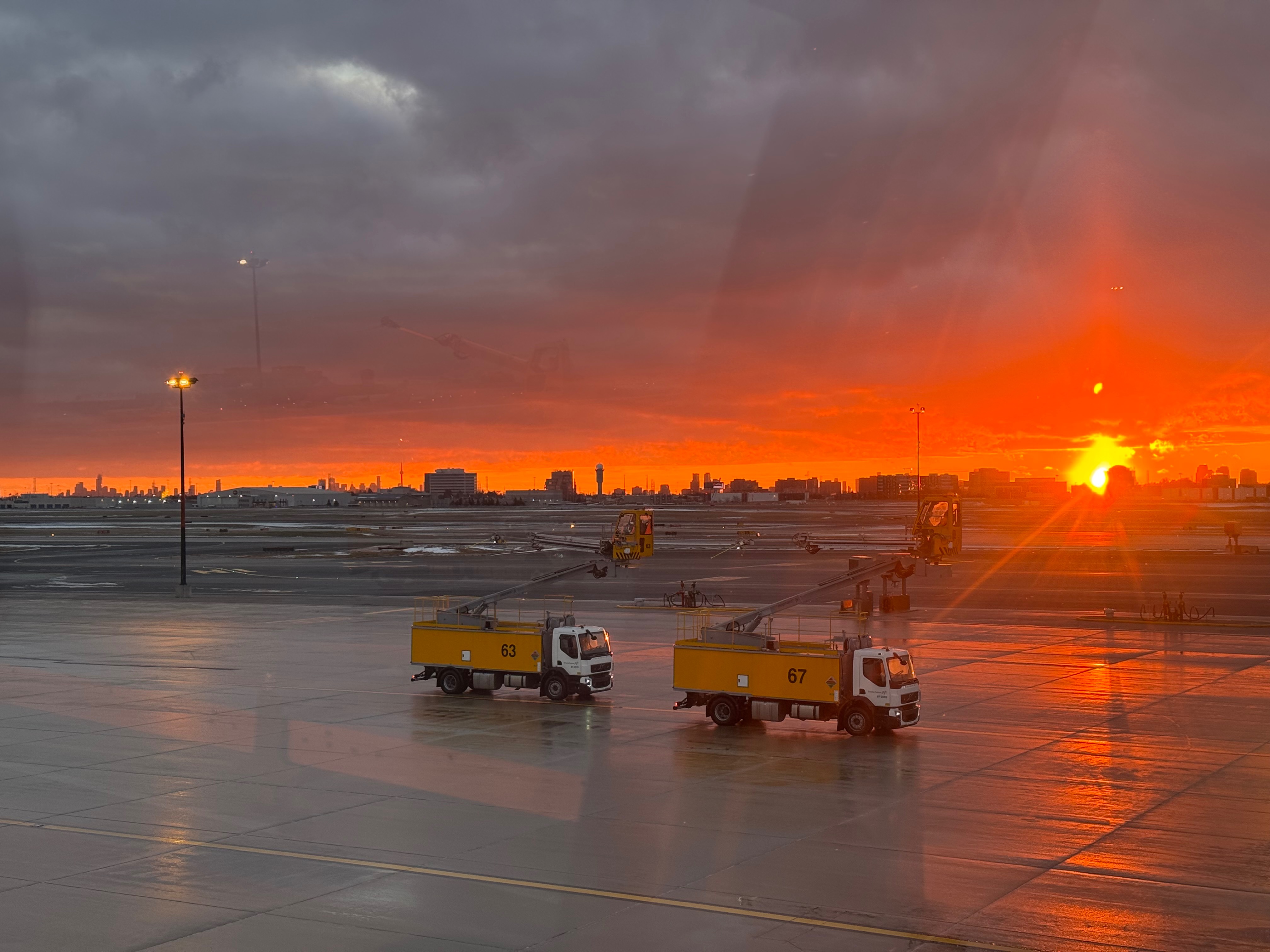 Sunset over an airport with orange and purple skies; two yellow service trucks labeled 63 and 67 on the tarmac.