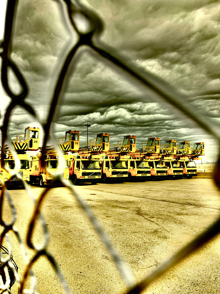 Yellow industrial vehicles numbered 12, 19, and 20 lined up behind a chain-link fence under a cloudy sky.