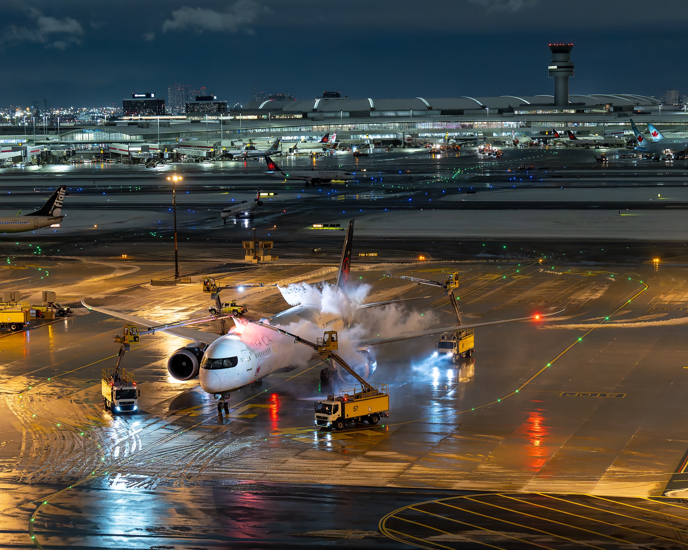 Aircraft being de-iced on a runway at night, with multiple de-icing vehicles and airport lights visible.