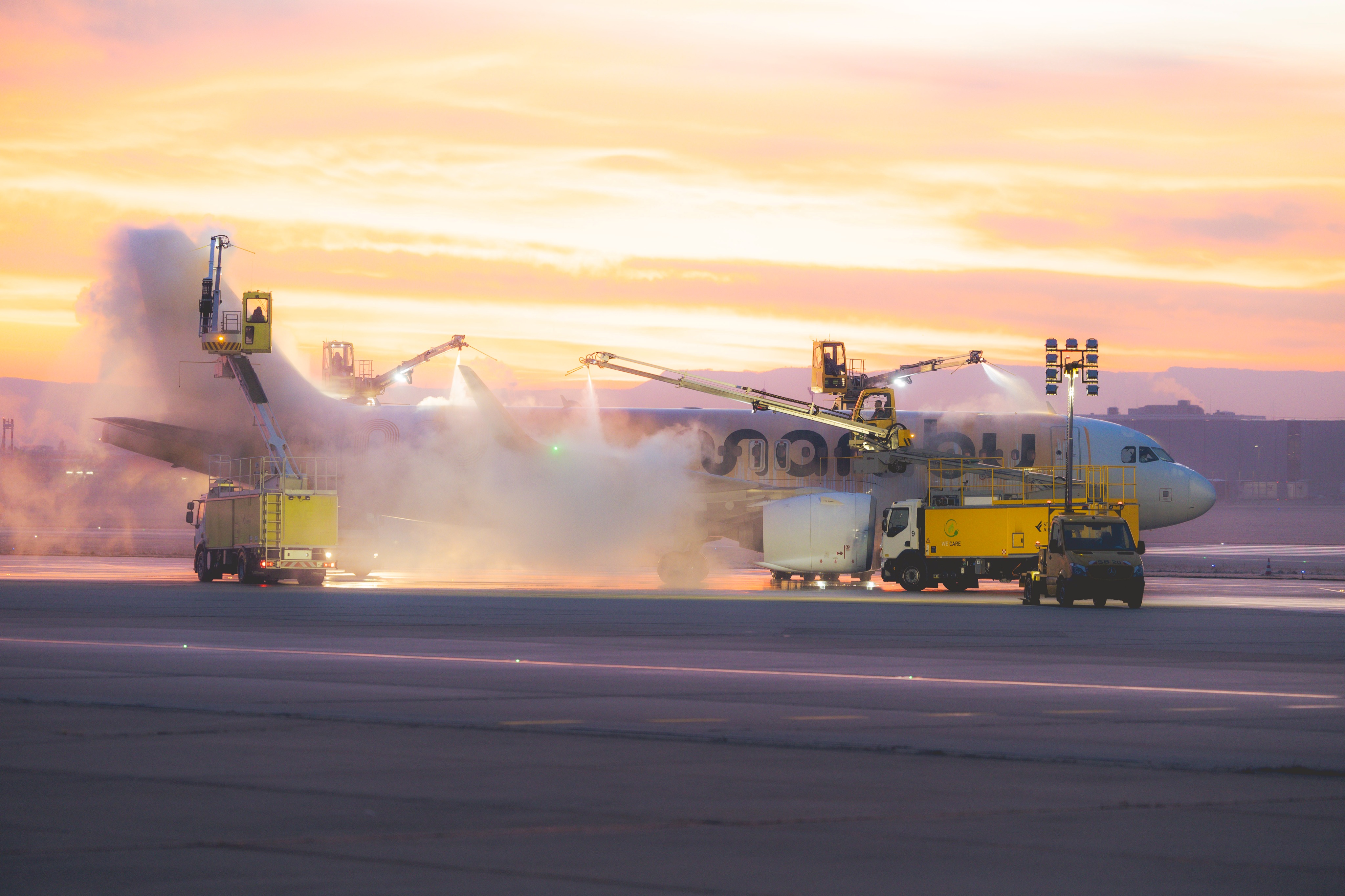Aircraft being serviced on the tarmac with water spray from two elevated platforms at sunset.