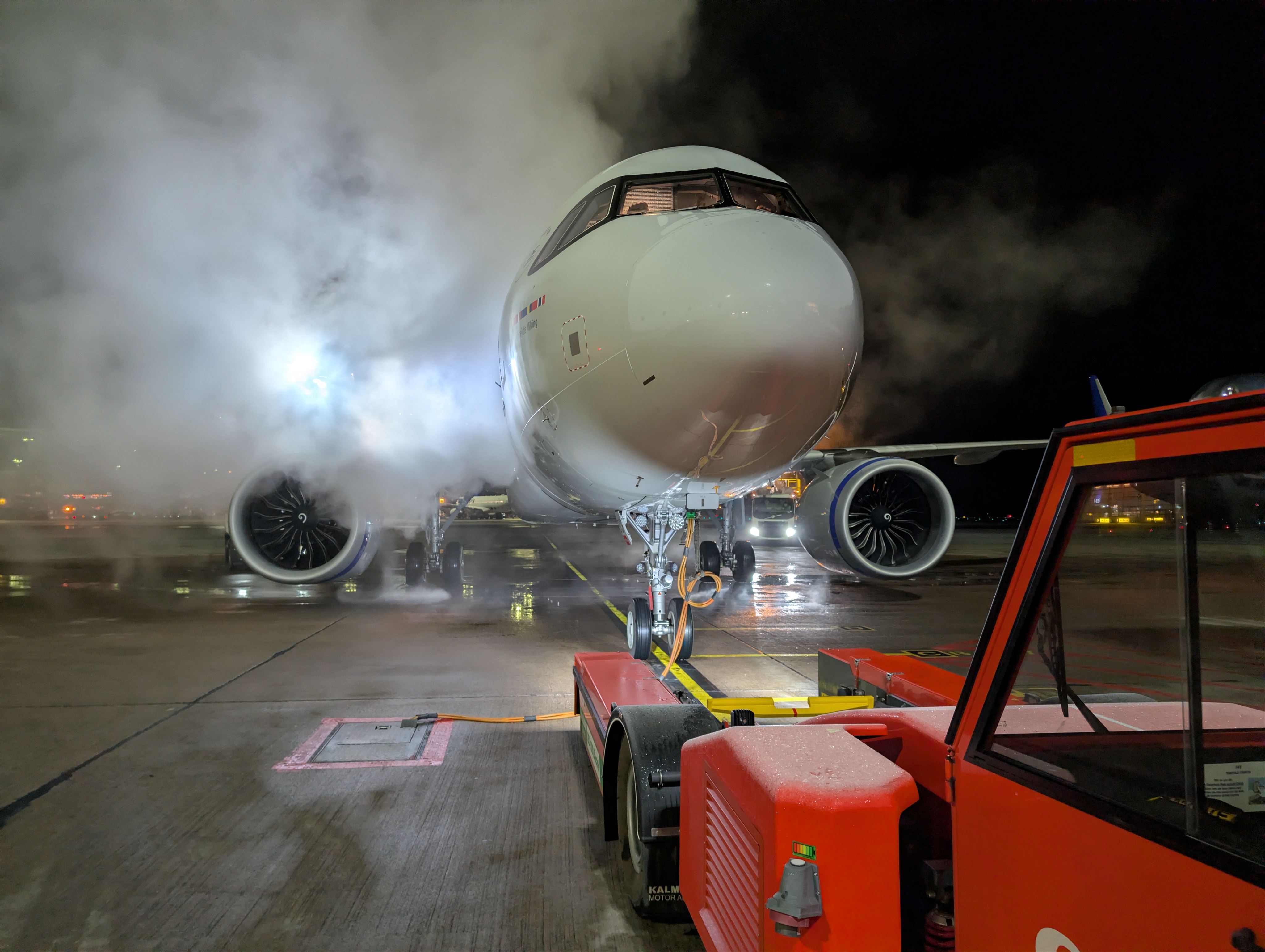 Airplane on tarmac with steam and lights at night, ground equipment visible in foreground.