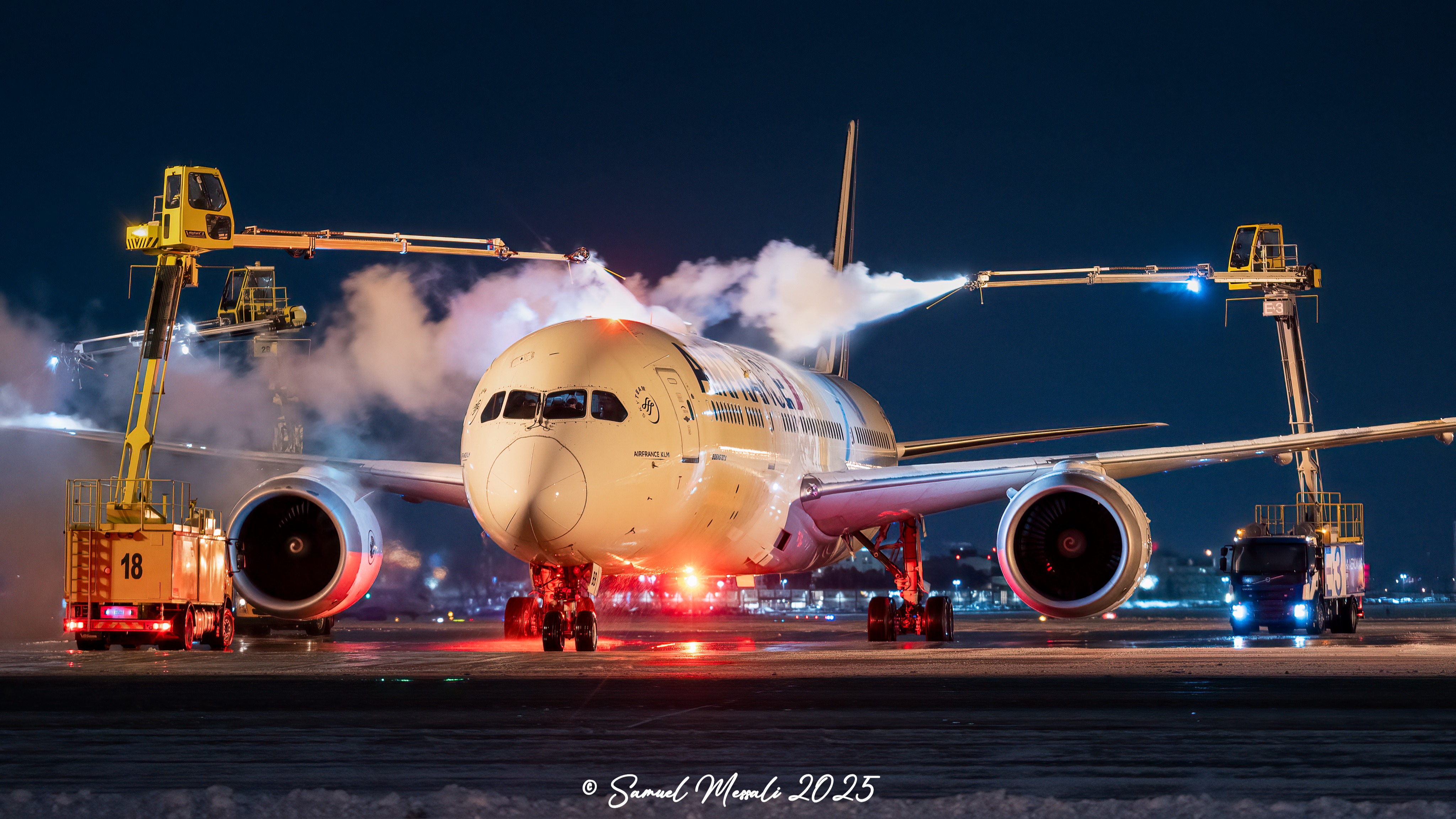 Boeing 787 aircraft being de-iced at night with two de-icing trucks, one labeled "18" and the other "43."