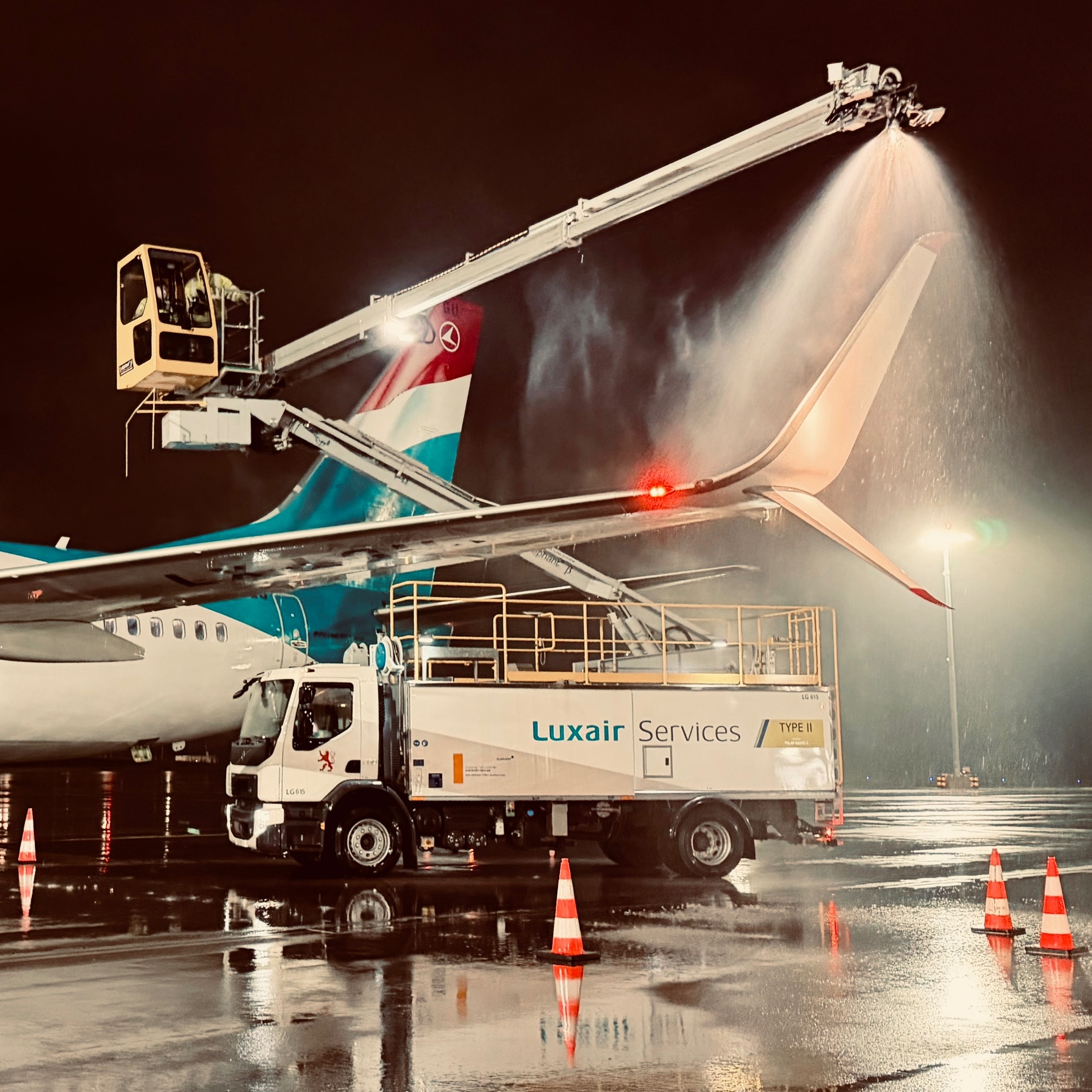Luxair Services truck spraying de-icing fluid on an airplane at night, with wet runway and safety cones visible.